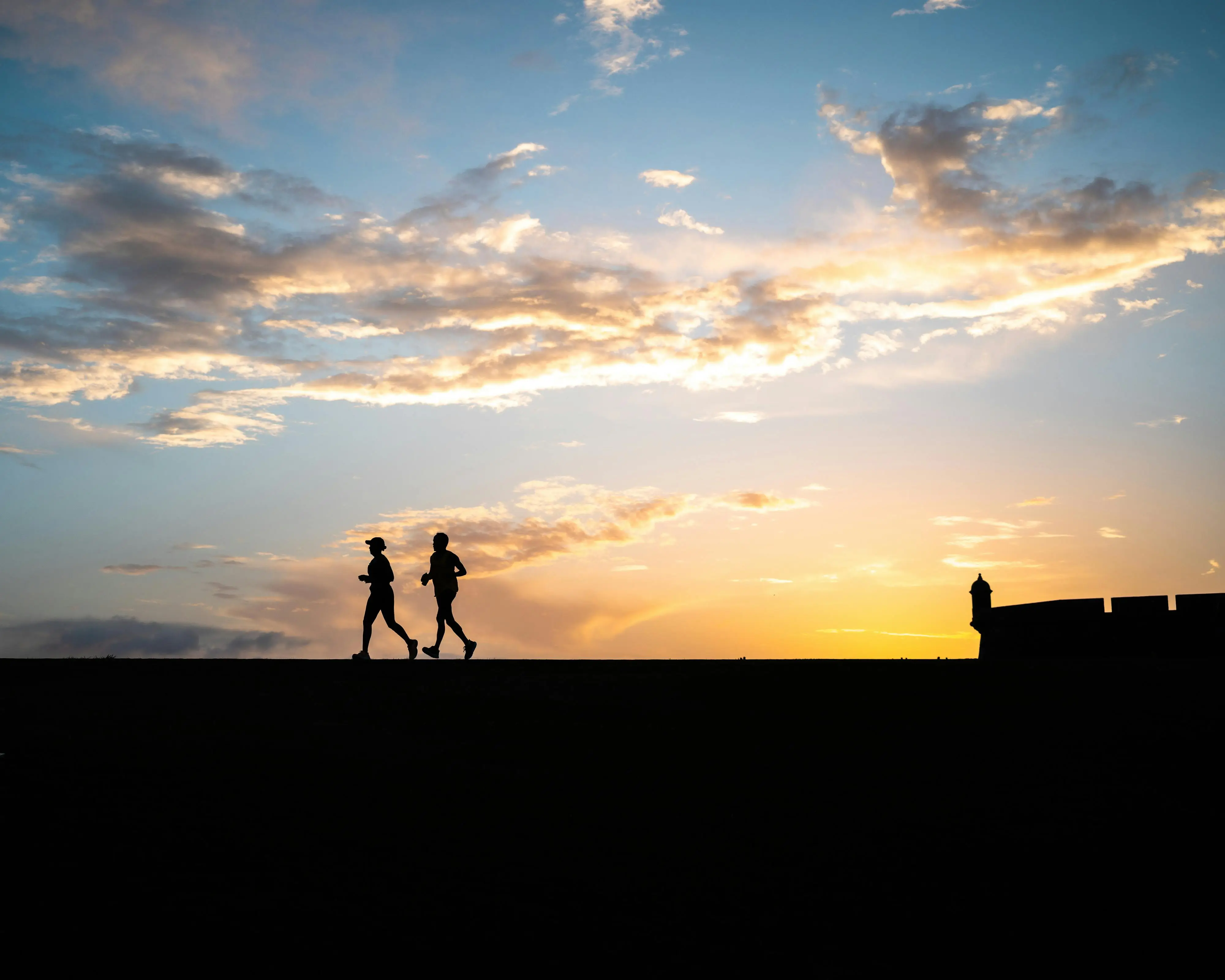 Two runners silhouetted against a sunset sky beside an old fortress wall