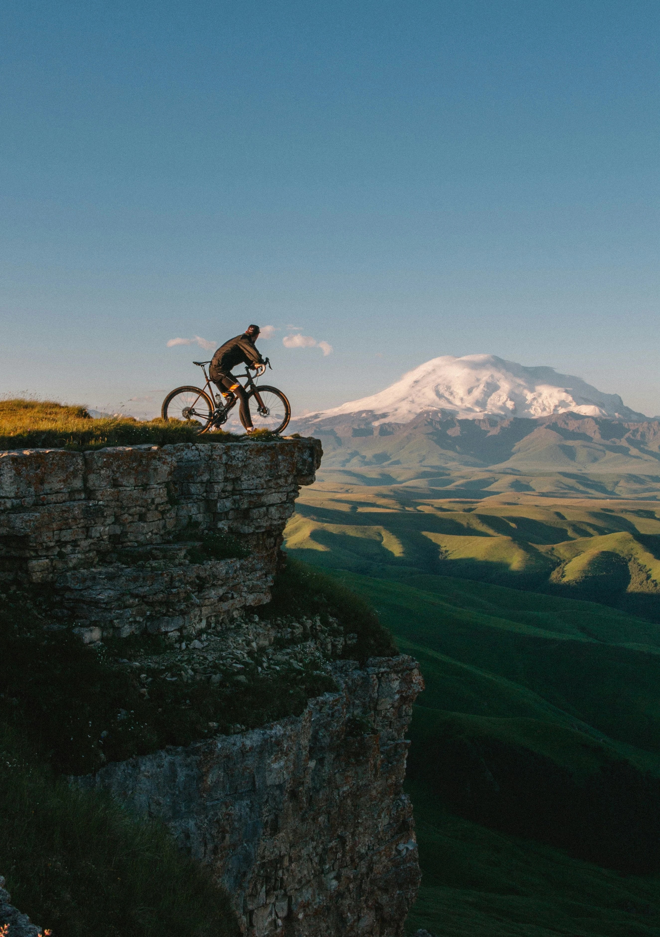 Cyclist with a road bike on a clifftop above green valleys and a snow-capped mountain