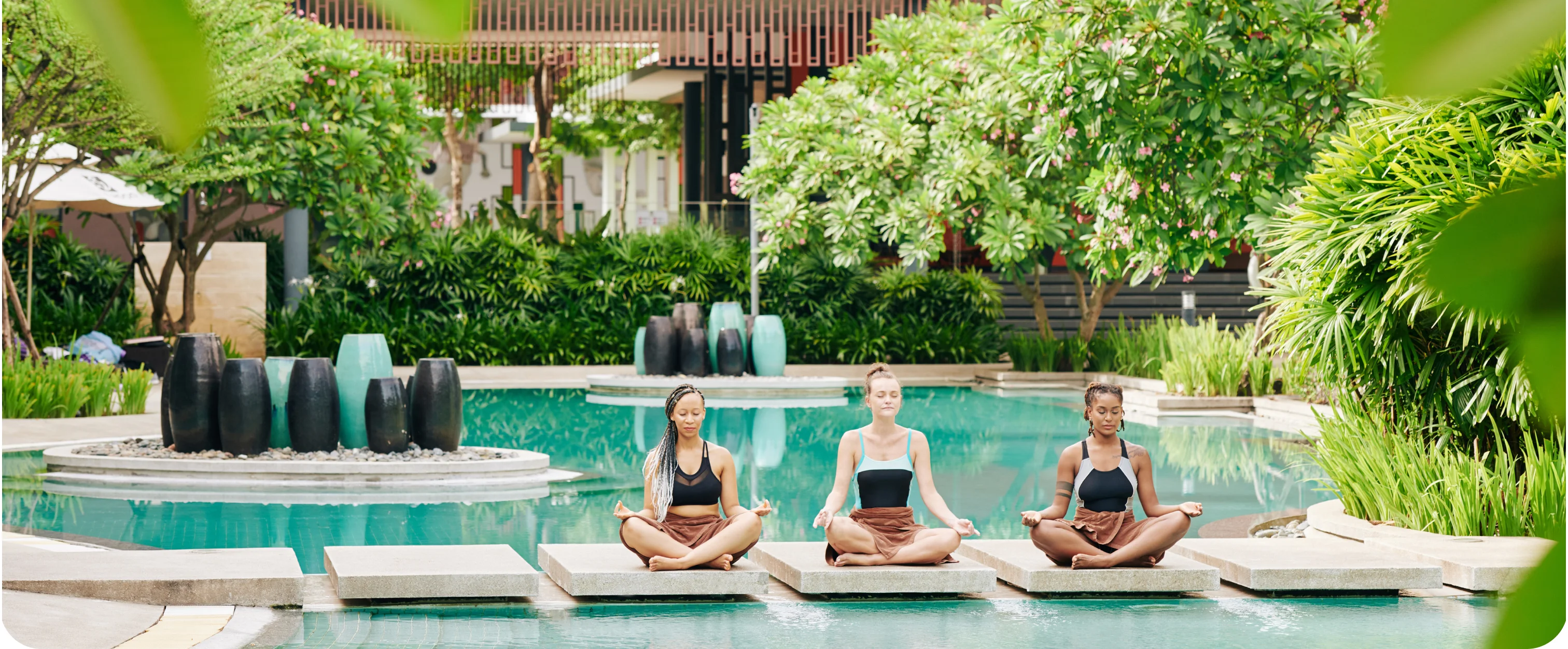 Three women meditating cross-legged beside a resort pool in a tropical garden